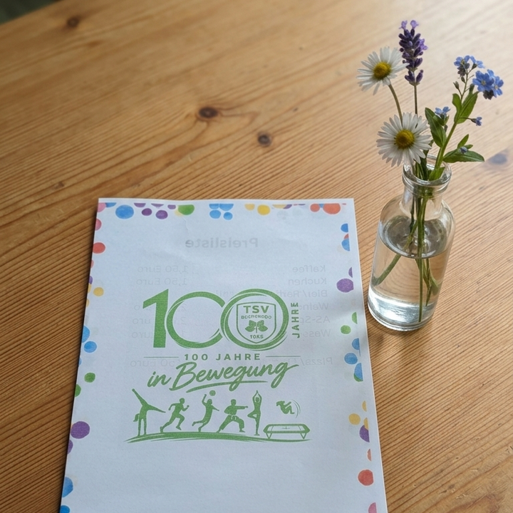 Auf einem Holztisch liegt ein weißes Faltblatt mit bunten Punkten am Rand. Darauf gedruckt ist das Logo des TSV Buchenrod, der grüne Schriftzug "100 JAHRE in Bewegung" sowie mehrere Silhouetten von Sportlern. Rechts neben dem Programmheft steht eine kleine Glasvase mit Wiesenblumen.