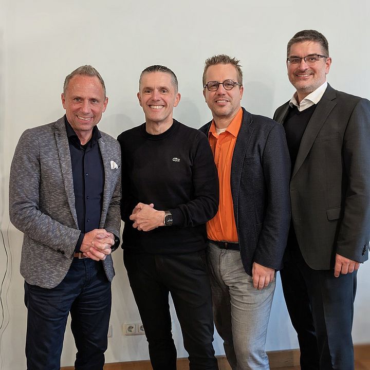 Vier stehende Männer in legerer Business-Kleidung blicken lächelnd nebeneinander in die Kamera vor einer schlichten weißen Wand.