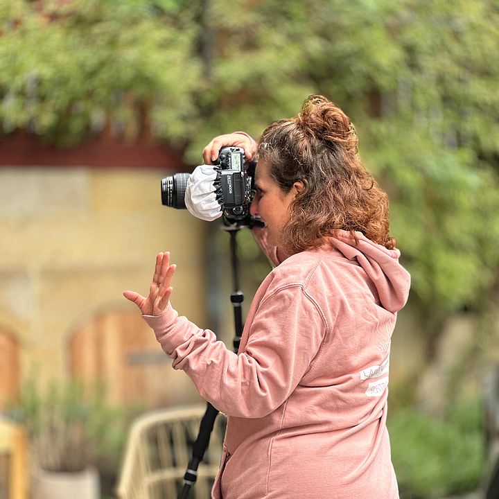  Eine Fotografin mit braunen, lockigen Haaren, die zu einem Dutt hochgesteckt sind, ist von der Seite zu sehen. Sie trägt einen rosa Kapuzenpullover und gestreifte Hosen. Sie schaut durch den Sucher einer Kamera, die auf einem Stativ montiert ist, und ihre linke Hand ist leicht erhoben, als würde sie Anweisungen geben oder gestikulieren. Der Hintergrund ist unscharf und zeigt Grünpflanzen sowie einen hellbraunen Stuhl mit einem hellblauen Kissen.