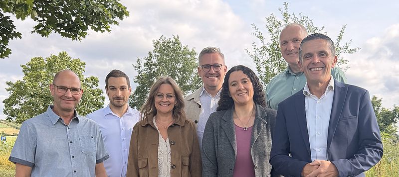 Sieben Personen, fünf Männer und zwei Frauen, stehen lächelnd nebeneinander auf einer sommerlichen Wiese. Die Gruppe trägt legere bis formelle Kleidung und posiert vor einem Hintergrund aus grünen Bäumen, einem Feld und blauem Himmel.